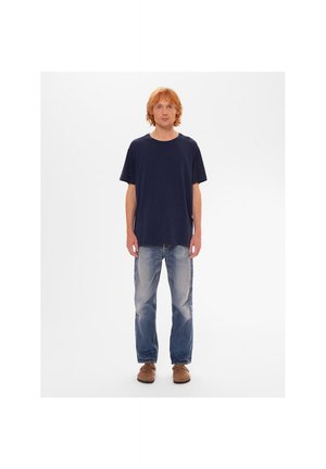 Hombre con cabello rojo de pie, vistiendo una camiseta azul marino, vaqueros azules descoloridos y zapatos marrones contra un fondo blanco liso.