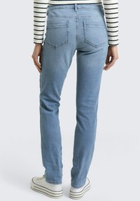 Osoba ubrana w jasnoniebieskie dżinsy i białe trampki z czarnymi paskami, stojąca tyłem do kamery na jednolitym tle.