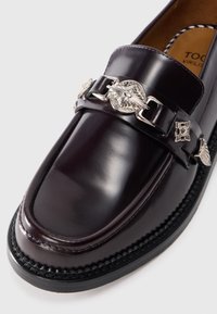 Mocasini din piele burgundă cu o cataramă decorativă argintie, talpă texturată neagră și finisaj neted. Design clasic cu accente împletite.