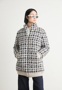 Puffjacke im Hahnentrittmuster mit hohem Kragen, Reißverschluss und gerippten Säumen. Farben sind beige, schwarz, braun und blassblau.