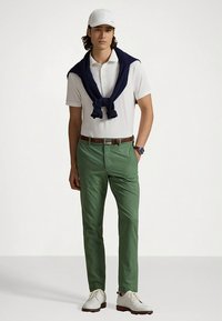 Vit pikétröja, marinblå tröja över axlarna, gröna byxor, brunt bälte och vita skor. Avslappnad outfit med rena linjer och färger.