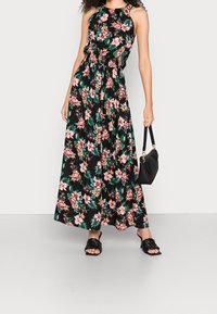 Frau mit einem ärmellosen, schwarzen Maxikleid mit pinkem Blumenmuster, die eine schwarze Handtasche hält und schwarze geflochtene Absatzsandalen trägt.
