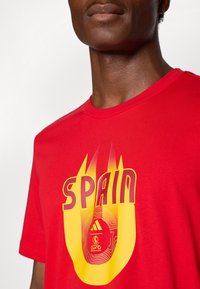 Rød bomulds t-shirt med et livligt gult og orange grafik med teksten "SPAIN" og et logo i midten. Korte ærmer og rund hals.