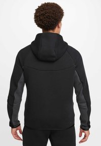 Sudadera con capucha negra y paneles laterales grises. Textura suave, ajuste atlético, con un cuello alto y una capucha estructurada. Branding mínimo visible.