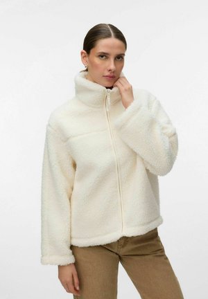 Žena s uhladenými vlasmi, oblečená v bielej flísovej bunde so zipsom a svetlohnedých nohaviciach, drží golier bundy jednou rukou.