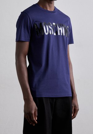 Mann trägt ein dunkelblaues kurzärmeliges T-Shirt mit glänzend schwarzem "MOSCHINO" Text über der Brust und schwarze Hose, stehend vor einem einfarbigen Hintergrund.
