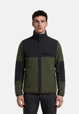 Young man wearing green and black zip-up jacket with high collar standing against plain background.