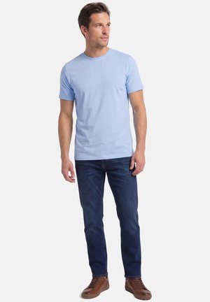 Mand iført lyseblå t-shirt med korte ærmer, mørkeblå jeans og brune sneakers, stående og kiggende til siden på hvid baggrund.