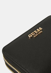 Portafoglio nero testurizzato con logo "GUESS" dorato in rilievo, dotato di chiusura con zip e dettagli di cucitura sottili.
