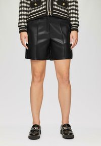 Zwarte leren shorts met een gestructureerd ontwerp, voorzien van voorplooien en een gladde textuur. Gecombineerd met glanzende zwarte loafers.