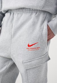 Šedé tepláky s elastickým pasem, bočními kapsami a náprsní kapsou. Obsahují červené logo Nike a text "NIKE ATHLETIC DEPT."