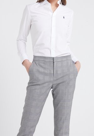 Persona con una camisa blanca ajustada de botones con un pequeño logo azul marino y pantalones de talle alto a cuadros grises, con las manos en los bolsillos.