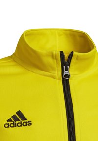Gelbe Polyesterjacke mit hohem Kragen, schwarzem Reißverschluss und schwarzem Adidas-Logo. Glatte Textur mit sportlichem Design.