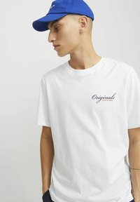 Bijela pamučna majica kratkih rukava sa tamnoplavi natpis "Originals Since 1990" na lijevoj strani prsnog koša. Uparena s plavim kapom.