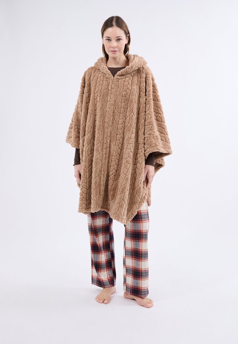 Hnědý, texturovaný fleecový pončo s kapucí, které má vzor pletené šňůry; nošeno přes kostkované pyžamové kalhoty v krémové, červené a černé barvě.