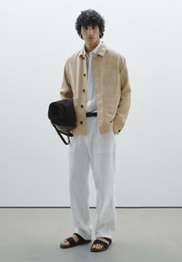 Veste beige clair à boutons avec une poche poitrine, associée à une chemise blanche et un pantalon blanc ample. Des sandales noires et un sac sombre complètent le look.