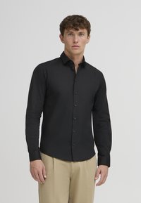 Uomo che indossa una camicia nera aderente con bottoni e pantaloni beige in piedi davanti a uno sfondo grigio chiaro uniforme.