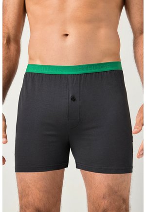 Torso masculino que lleva calzoncillos tipo bóxer negros con una cinturilla elástica verde y un solo botón negro en el centro delantero.