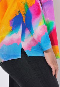 Felle multicolore lange mouwen shirt met abstract aquarelpatroon, gedragen met zwarte broek, hand rustend op de dij.