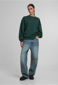 Zelená nadrozmerná mikina so spadnutými plecami, kombinovaná s svetlomodrými voľnými džínami. Čierne robustné topánky dopĺňajú outfit. Hladká textúra.