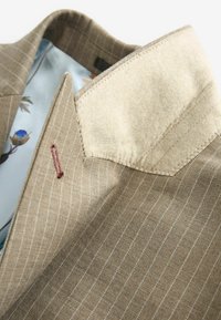 Brauner Nadelstreifen-Blazer mit einem beigen Filzkragen, ausgestattet mit einem Futter mit Blumenmuster und roten Stickdetails an der Seite.