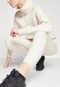 Costum de sport bej deschis, cu guler înalt și fermoar, fabricat dintr-un material moale, purtat cu pantofi sport negri, cu șireturi și accente texturate.