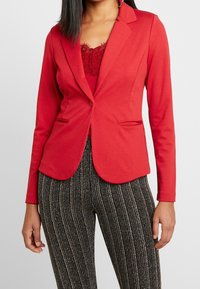 Blazer stretch rouge à coupe ajustée, avec revers crantés et deux poches avant, associé à un pantalon noir texturé à motifs.
