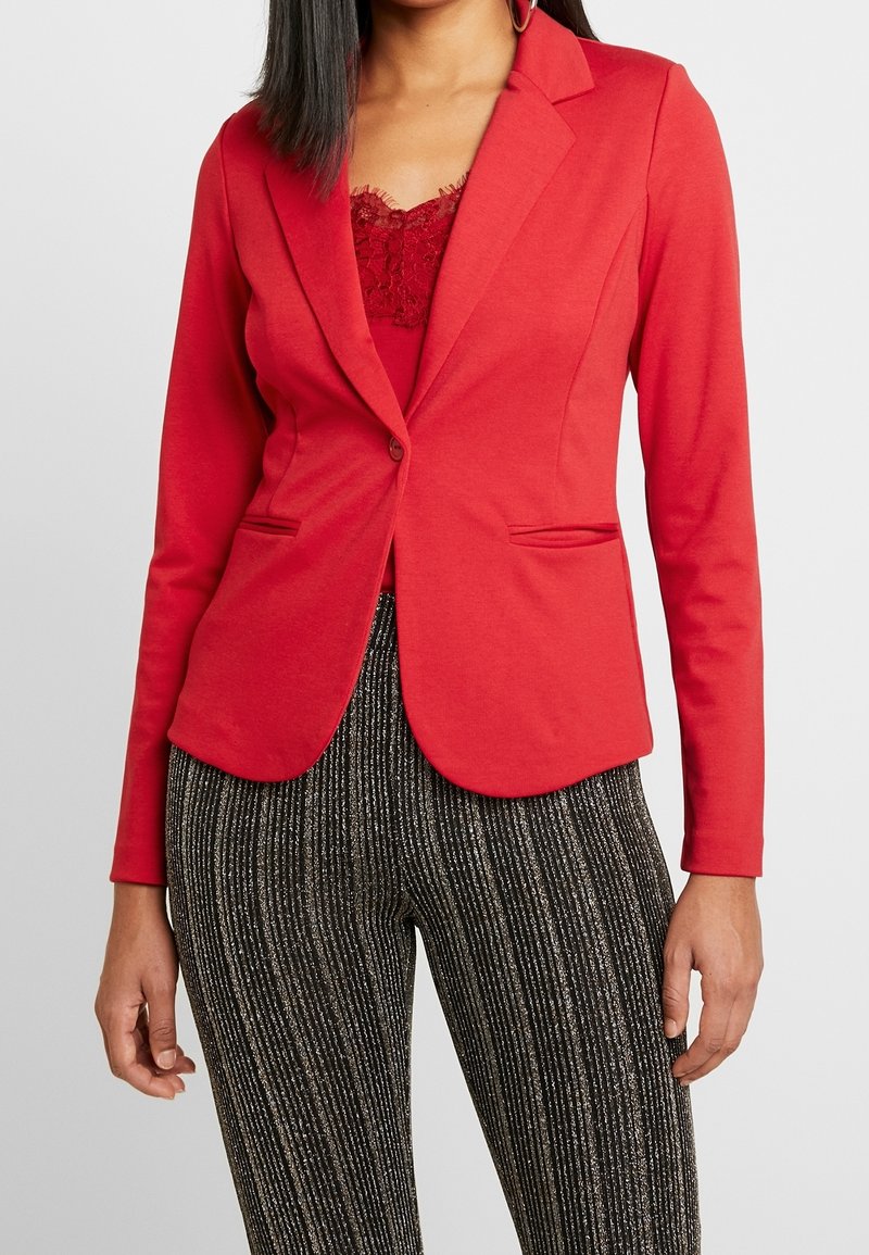 Blazer stretch rouge à coupe ajustée, avec revers crantés et deux poches avant, associé à un pantalon noir texturé à motifs.