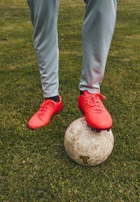 Røde fotballsko med teksturerte overflater og lisser, står på en slitt grå fotball, på gress med synlige strå.
