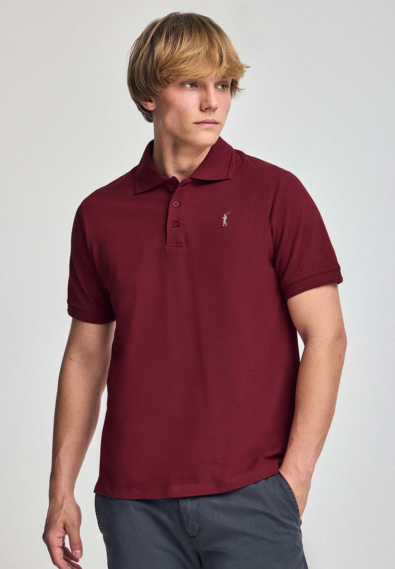 Bordeauxfärgad polo t-shirt i bomullsblandning med tvåknappsstängning, korta ärmar och en liten logotyp på bröstet. Slät textur.