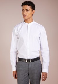 Camisa de algodón blanca con cuello tipo mao, con botones en el frente y mangas largas, combinada con pantalones de cuadro gris y un cinturón negro.