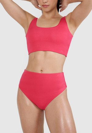 Kobieta nosząca teksturowane, jasno różowe majtki bikini z wysokim stanem i dopasowany krótki top z rękami uniesionymi za głową na jednolitym tle.