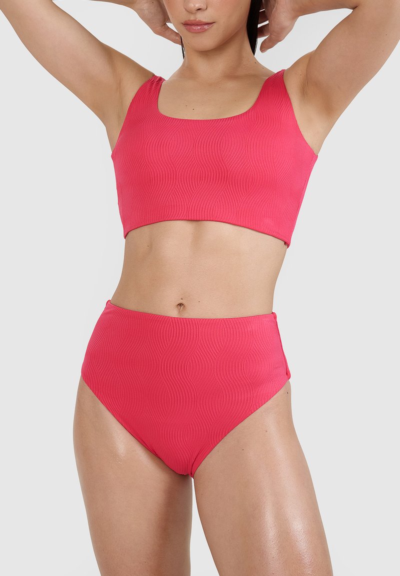 Femme portant un bas de bikini taille haute rose vif texturé et un crop top assorti, bras levés derrière la tête sur fond uni.