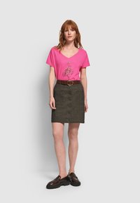 Mujer con cabello rojo que lleva una camiseta de cuello en V rosa con un diseño de flamenco, cinturón marrón, falda oscura abotonada y zapatos oscuros tipo slip-on.