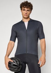 Cyklistický dres s krátkym rukávom v tmavošedej farbe s vysokým golierom a predným zipsom. Model drží čiernu prilbu, v kombinácii s čiernymi cyklistickými šortkami.