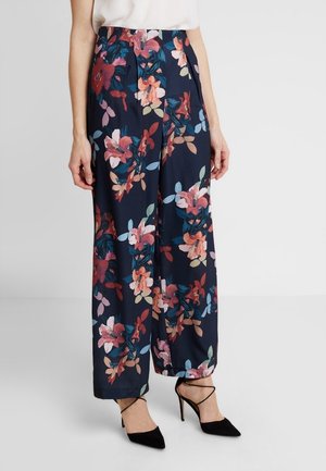 Pantalon ample bleu marine à imprimé floral multicolore, porté avec un haut blanc et des talons noirs à bout pointu avec brides à la cheville.