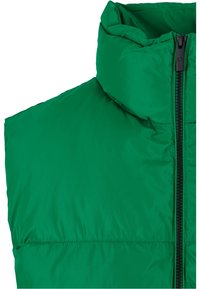 Grön pufferväst med hög krage, med en central svart dragkedja och en slät, glänsande tygstruktur. Lätt och isolerad design.
