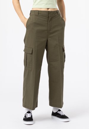 Person, die olivgrüne Cargohosen mit Seitentaschen, weiße Socken und schwarze Sneakers trägt, steht mit Händen in den Taschen vor weißem Hintergrund.