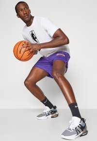 Korvpallur, kes kannab valget T-särki, lilac-lühikesi pükse, millel on "Lakers" logo, ja mustade-valgete tossude, hoiab käes korvpalli.