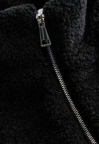 Zwarte fleece stof met een zachte, getextureerde uitstraling, voorzien van een zilveren ritssluiting aan de voorkant. Duurzaam en warm ontwerp.