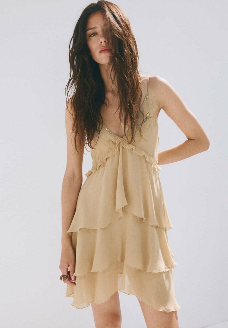 Femme aux longs cheveux foncés portant une robe beige sans manches à plusieurs couches, debout devant un arrière-plan clair uni.