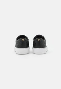 Schwarze, strukturierte Slipper mit einer weißen Sohle, die an der Rückseite ein kleines goldfarbenes Logo-Detail und ein glattes Innenfutter aufweisen.