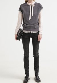 Sweat-shirt gris à col avec un nœud papillon, porté sur une chemise rose clair à pois. Porté avec un jean skinny noir et des chaussures noires, tenant une pochette.