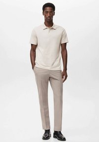 Camisa polo beige de mangas cortas, combinada con pantalones de color marrón claro. Zapatos negros, de diseño liso, sin patrones ni acentos visibles.