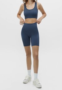 Modrý sportovní top na tělo a vysoké šortky, vyrobené z hladkého materiálu. Modelka je doplnila světle barevnými teniskami a sportovními ponožkami.