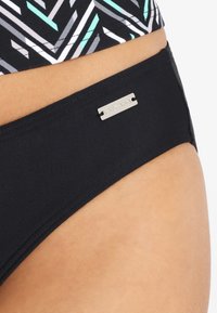 Zwarte bikini-broek met een gladde textuur, met een zilveren merkembleem. De achtergrond is gedecoreerd met zwarte, witte en aqua geometrische vormen.