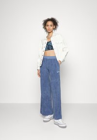 Calças de veludo azul de perna larga com uma cintura alta, combinadas com um top cropped estampado em preto e azul, e uma jaqueta de ganga branca. Ténis brancos.