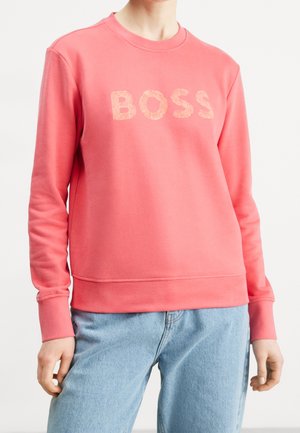 Sudadera color rosa coral con el logo "BOSS" en letras texturizadas, cuello redondo, puños y dobladillo acanalados. Combinada con jeans azul claros.
