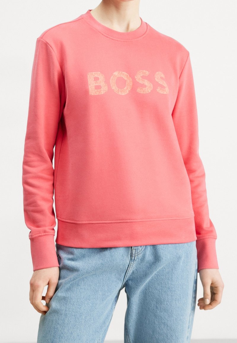 Sudadera color rosa coral con el logo "BOSS" en letras texturizadas, cuello redondo, puños y dobladillo acanalados. Combinada con jeans azul claros.