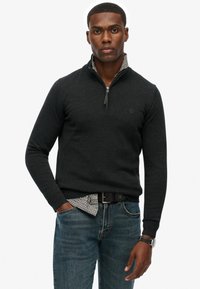 Donkergrijze sweater met rits en geribbelde manchetten, gedragen over een geblokt shirt. Gecombineerd met blauwe jeans, met een zwarte riem en horloge.